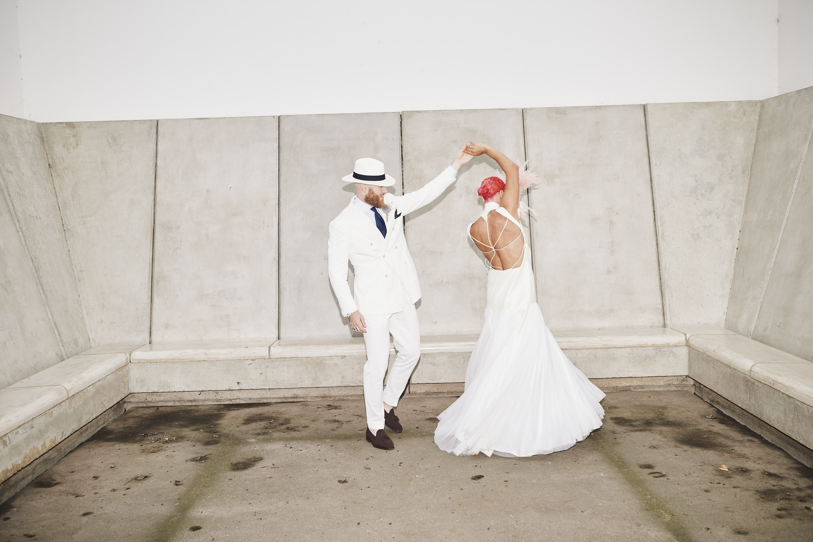 A bride and groom, wearing a long white dress and white suit, dancing inside a small room with concrete seating.