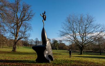 A man looking at a sculpture of a hare balanced on the tip of an oversized anvil, displayed outdoors.