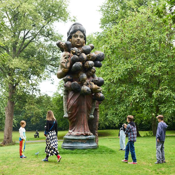 A large statue of a woman covered in small heads, stands in a park. Four children, two boys and two girls, explore around the statue, surrounded by lush green trees and grass. The atmosphere is serene and playful.