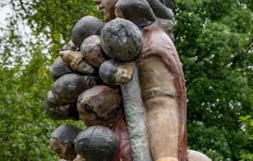 A large, intricately detailed sculpture of a woman holding multiple small figures, set against a backdrop of lush green trees. The woman has a serene expression, and the figures appear to be clinging to her.