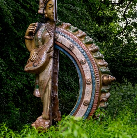 A sculpture of a deity with stands amidst lush greenery, surrounded by tall trees.