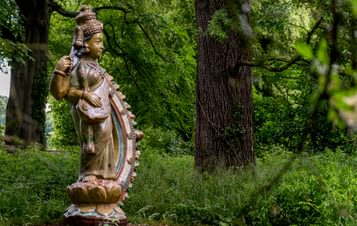 A sculpture of a deity with stands amidst lush greenery, surrounded by tall trees.
