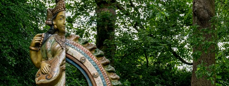 A sculpture of a deity with stands amidst lush greenery, surrounded by tall trees.