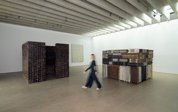 A blurred figure walking between two square installations, one made from bricks, one made from stacked radiators, in a gallery space.