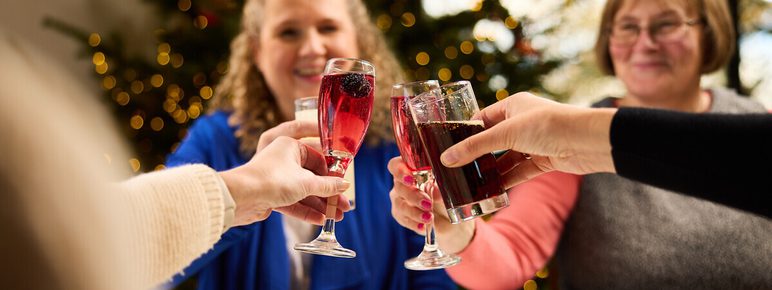 A festive gathering featuring three people toasting with drinks at a table adorned with food. In the background, a decorated Christmas tree glimmers with lights.