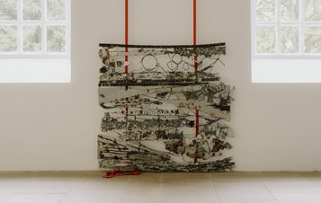 A welded metal artwork with abstract shapes, suspended from red straps against a white gallery wall, with tall windows to either side.