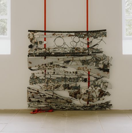 A welded metal artwork with abstract shapes, suspended from red straps against a white gallery wall, with tall windows to either side.