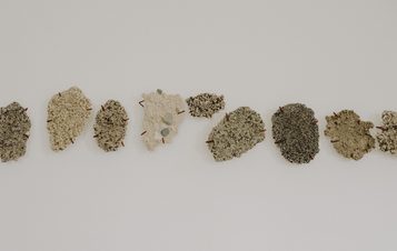 A row of textured artworks in muted shades embedded with stones and sand, displayed on a white wall.