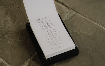 Detail of a suspended paper work with hand written poetry inside. It's displayed resting on a black plinth at floor level, on a stone floor.