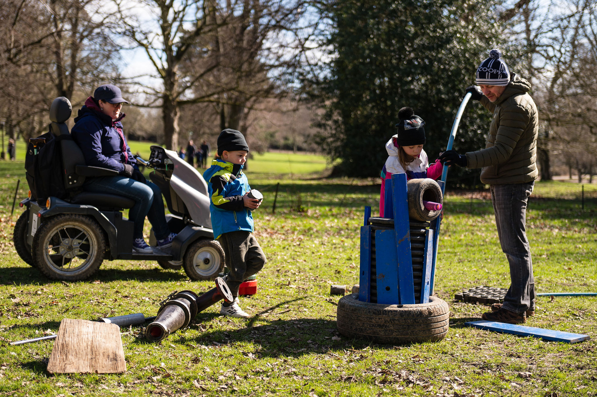 A family of 2 adults and 2 children building sculptures out of typers and plastic pipes. One of the adults is using a tramper mobility scooter.