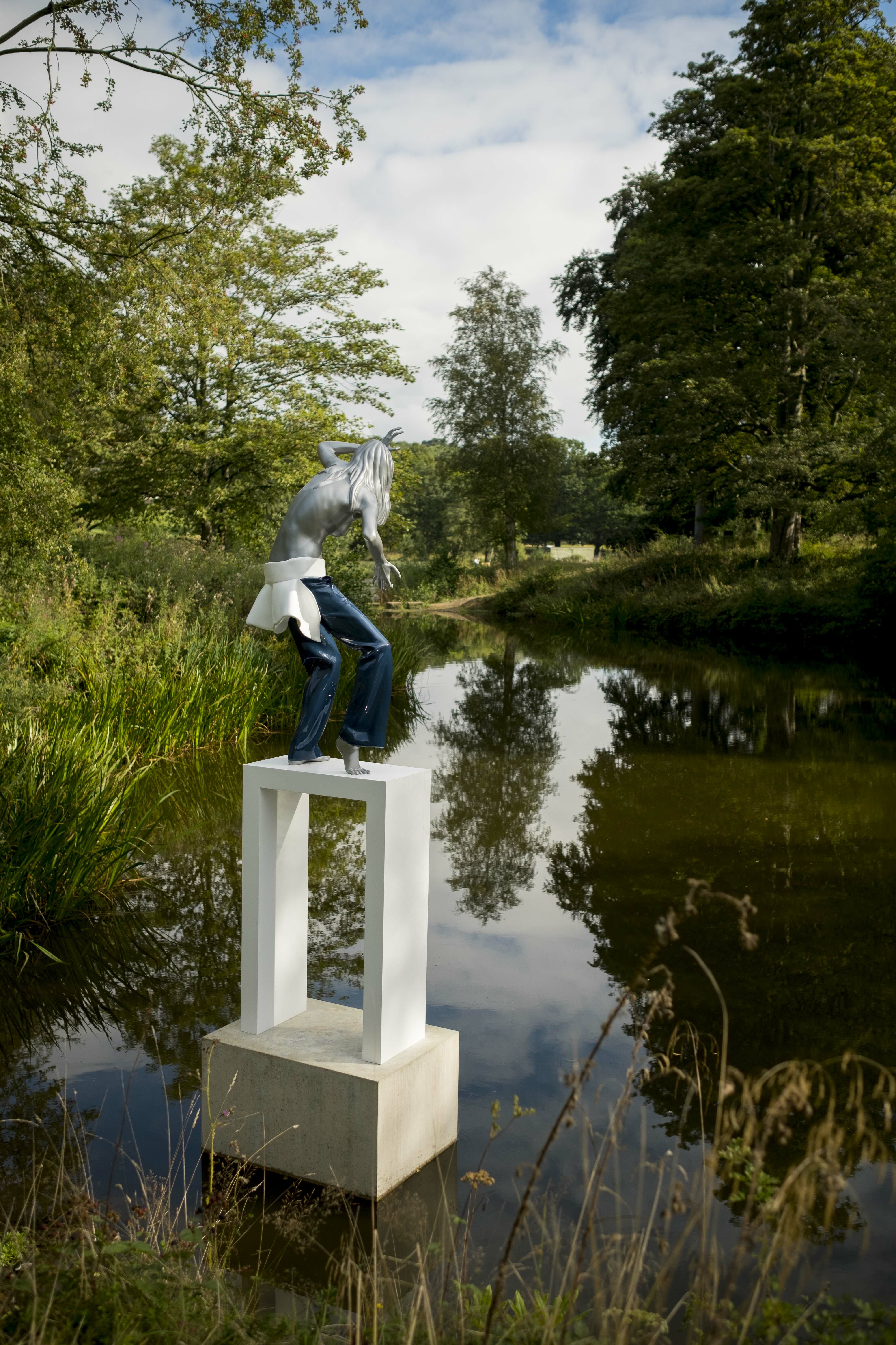 A female figure wearing jeans standing on a white plinth at the edge of a lake, surrounded by foliage