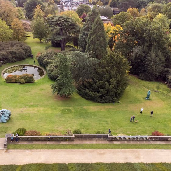 An aerial photograph of a formal garden with large trees, bronze classical sculptures and people.