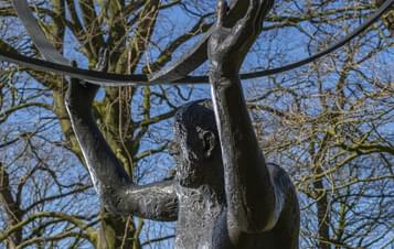 A bronze statue of a muscular man lifting a barbell overhead, set against a backdrop of bare trees and a clear blue sky. The figure's expression conveys determination, highlighting the theme of strength and perseverance.