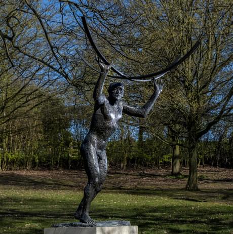 A bronze sculpture of a slender figure holding a large curved object above their head, set in a grassy area surrounded by trees. The sky is clear and blue, creating a serene atmosphere in the park.