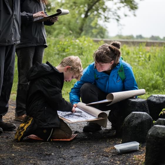 An adult and child crouching near to black lump-like objects while drawing onto clipboards and paper.