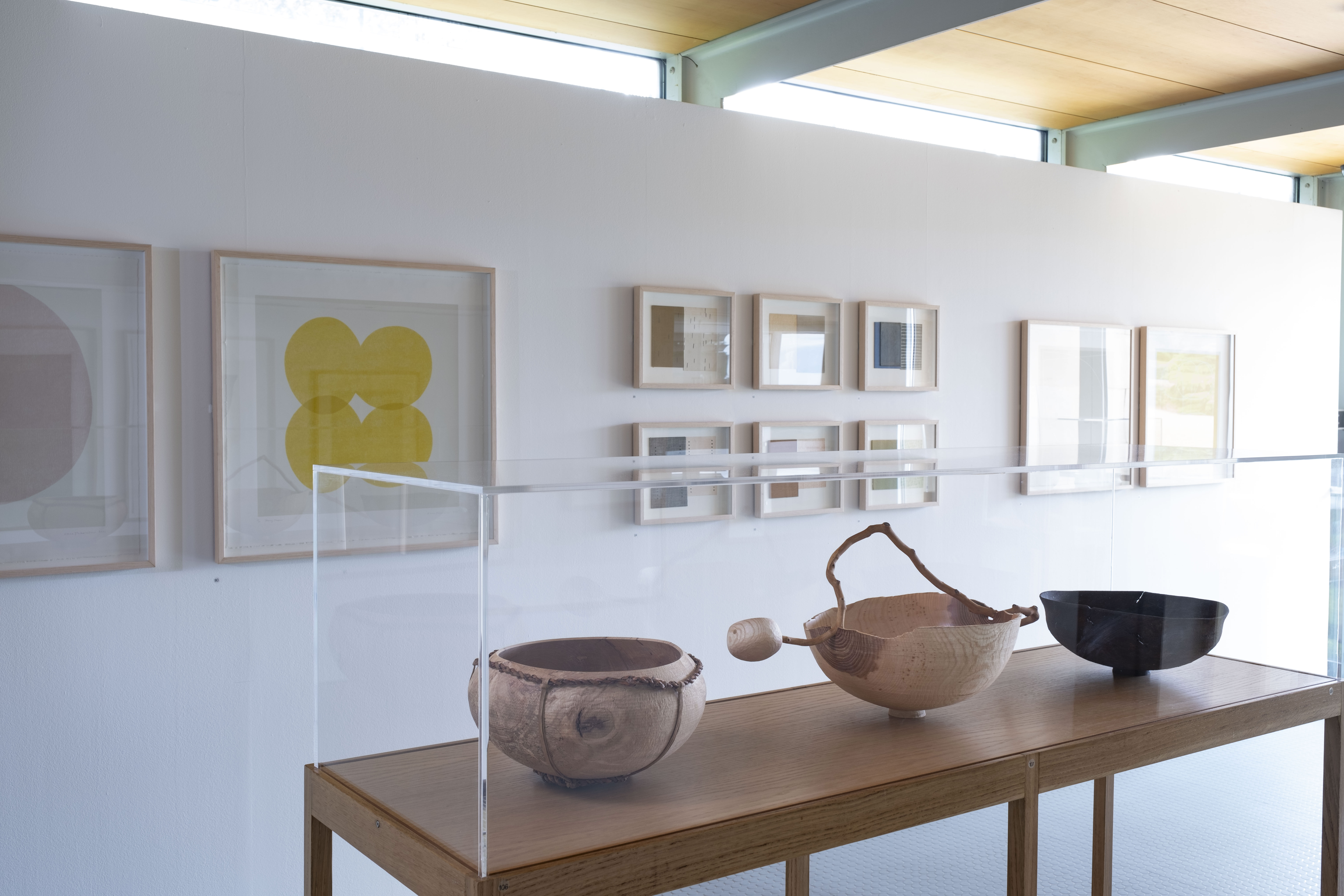 A white gallery space with framed prints on the walls and carved wooden vessels on a table in the centre