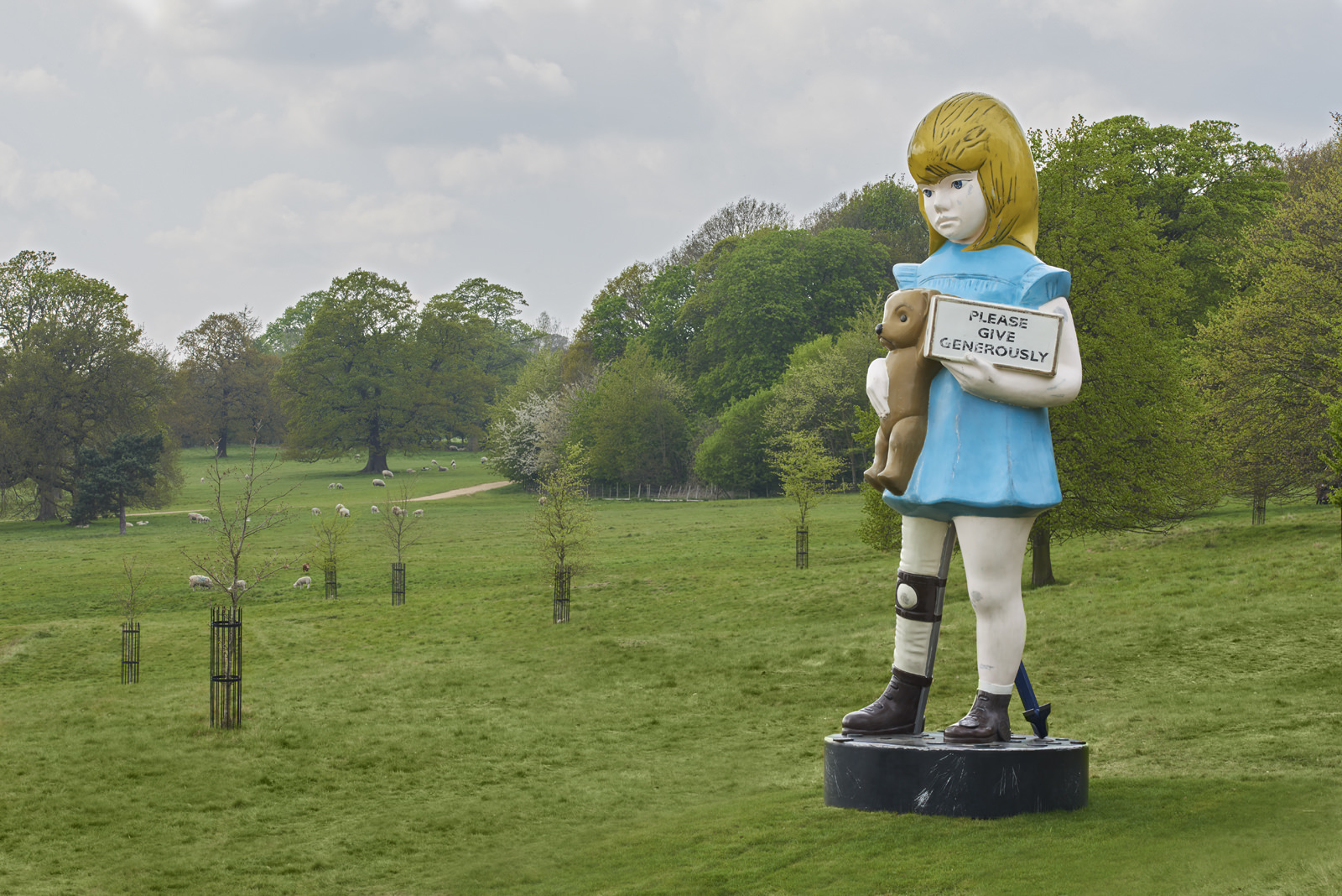 Sculpture of a young girl holding a teddy bear and a charity box, among saplings in YSP parkland.