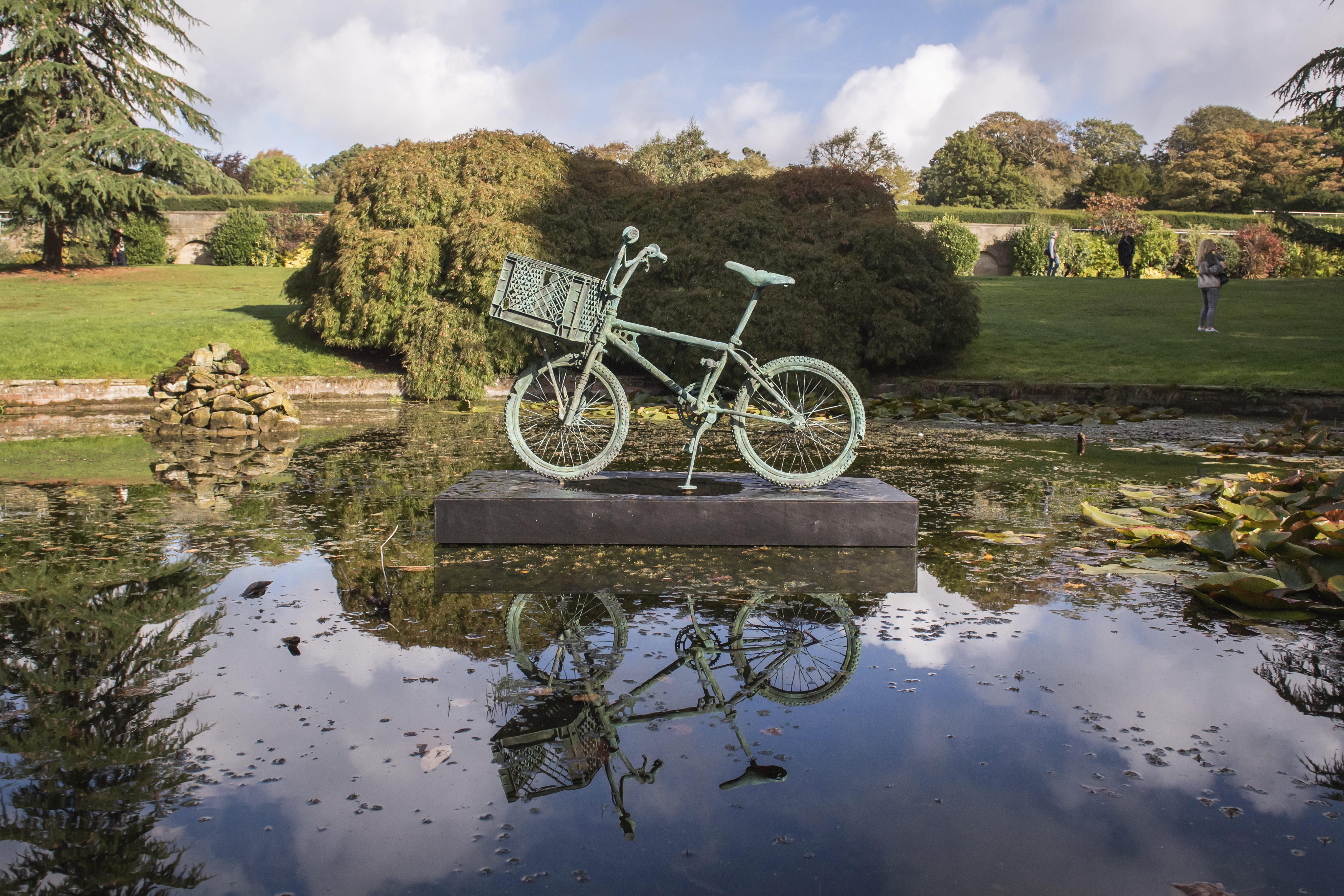 A bronze bicycle mounted on a plinth in the centre of a pond, with bushes and trees in the background.