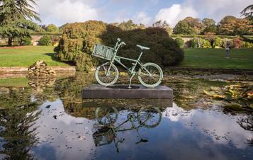 A bronze bicycle mounted on a plinth in the centre of a pond, with bushes and trees in the background.