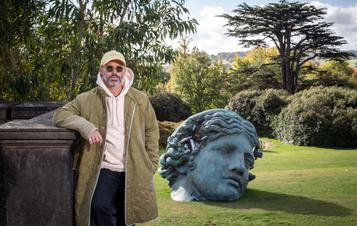 Daniel Arsham, a white man wearing sunglasses and a khaki coat, standing in front of a large bronze sculpture of a woman's head, leaning on a wall.