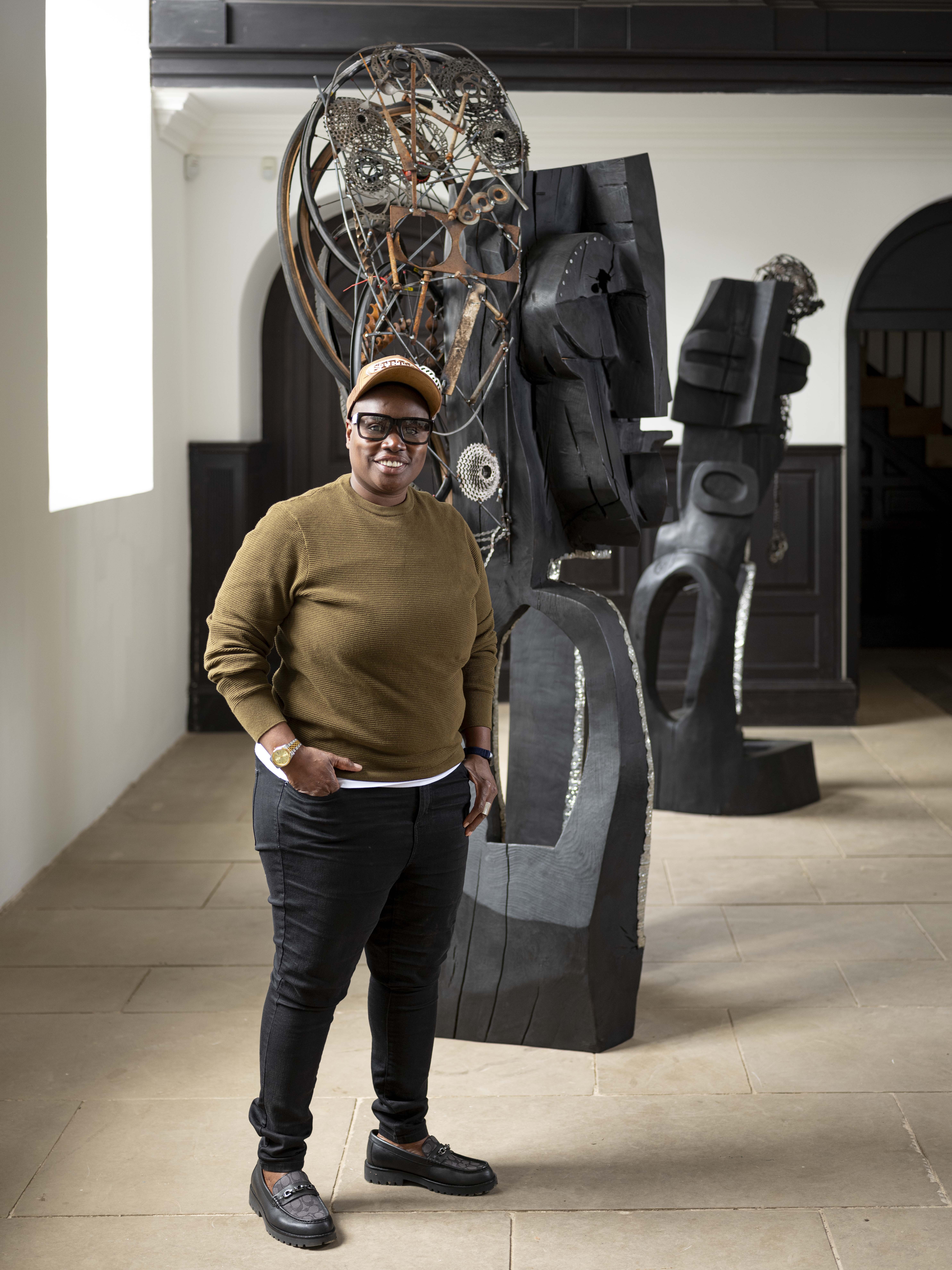 A black woman wearing a cap and glasses, stands in front of a carved wooden figure with her hands in her pockets.
