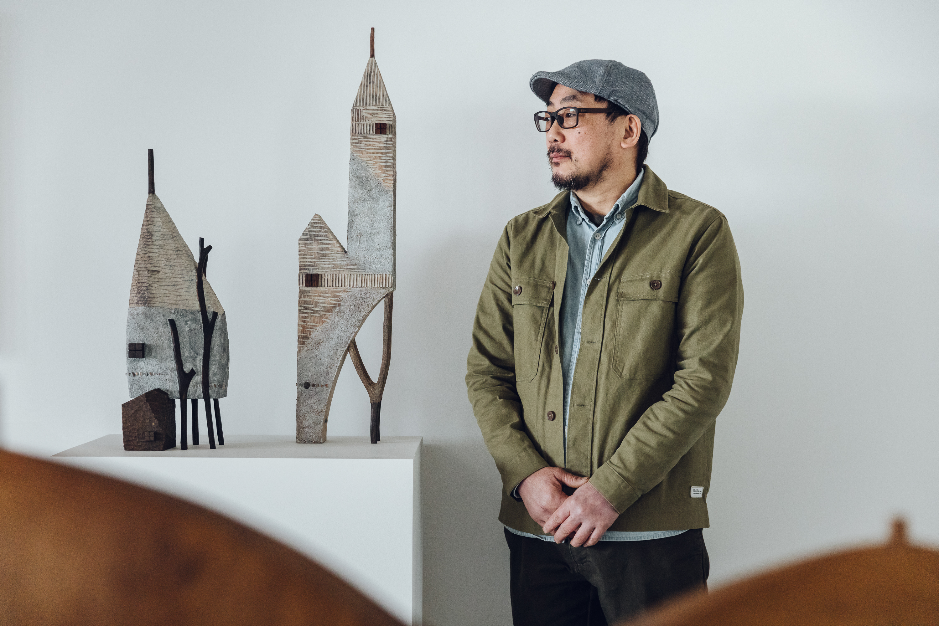 The artist Yukihiro Akama, a Japanese man wearing a green jacket and grey cap, standing next to two carved wooden houses.