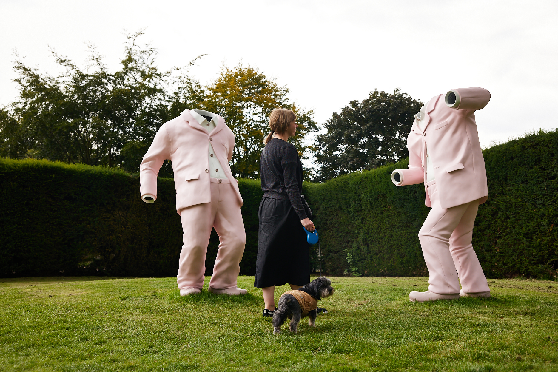 A woman walking a small dog looking at two giant pink suit sculptures.