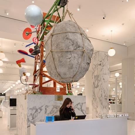 A colourful scaffolding structure with a giant boulder suspended over a person sitting at a desk.