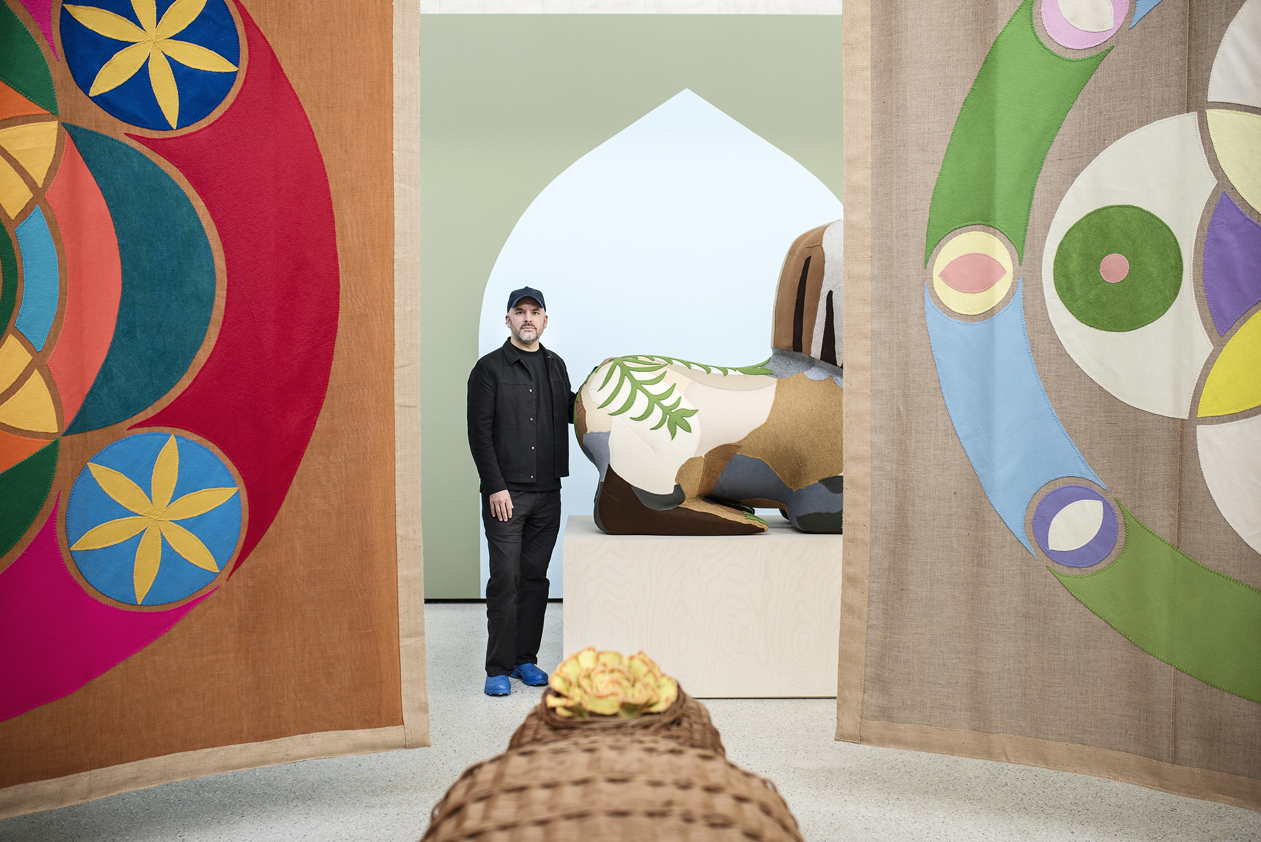 A man wearing black trousers and jacket, standing between two colourful fabric wall hangings in a gallery space.