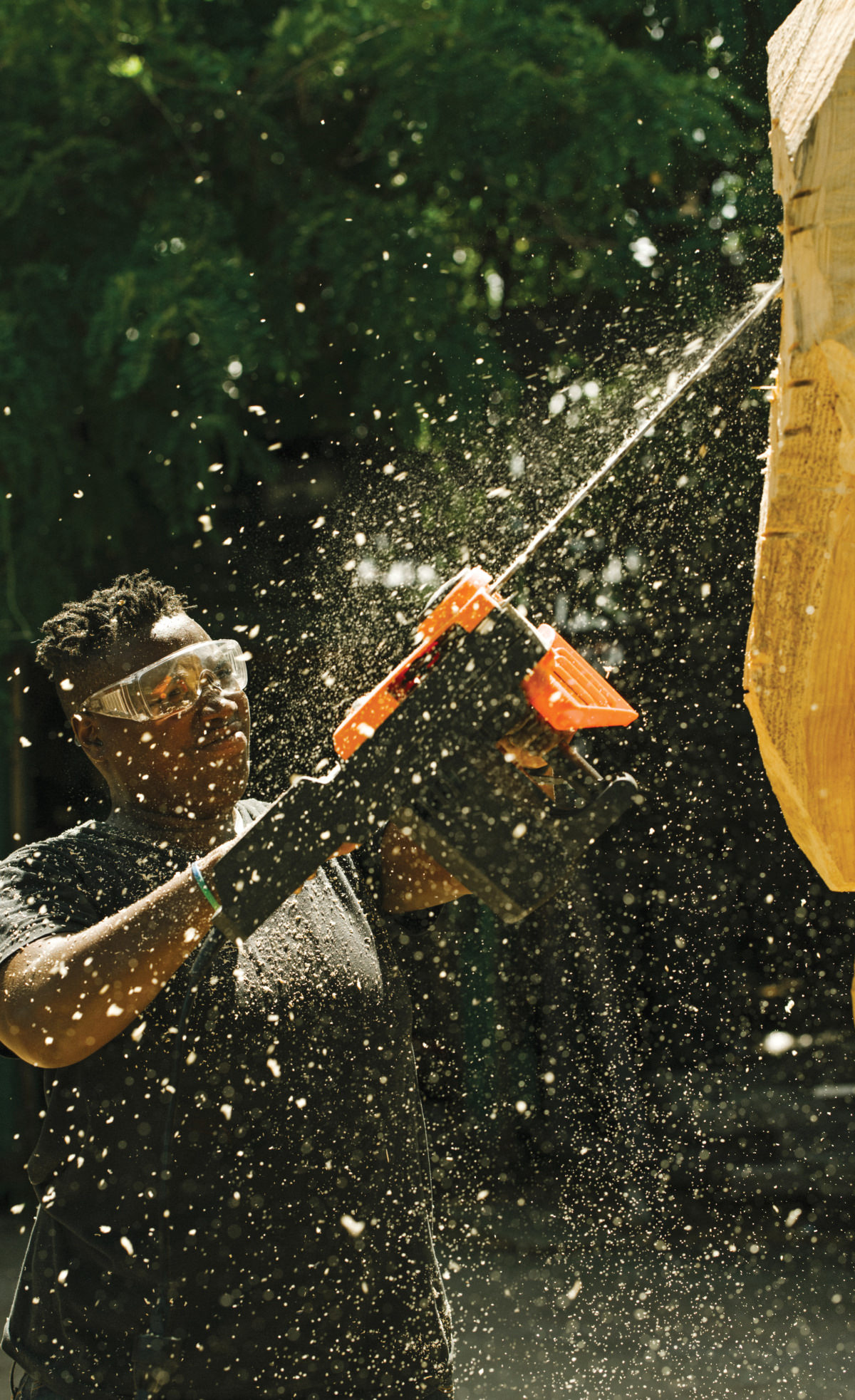 A black woman wearing safety goggles, carving a piece of wood using a chainsaw
