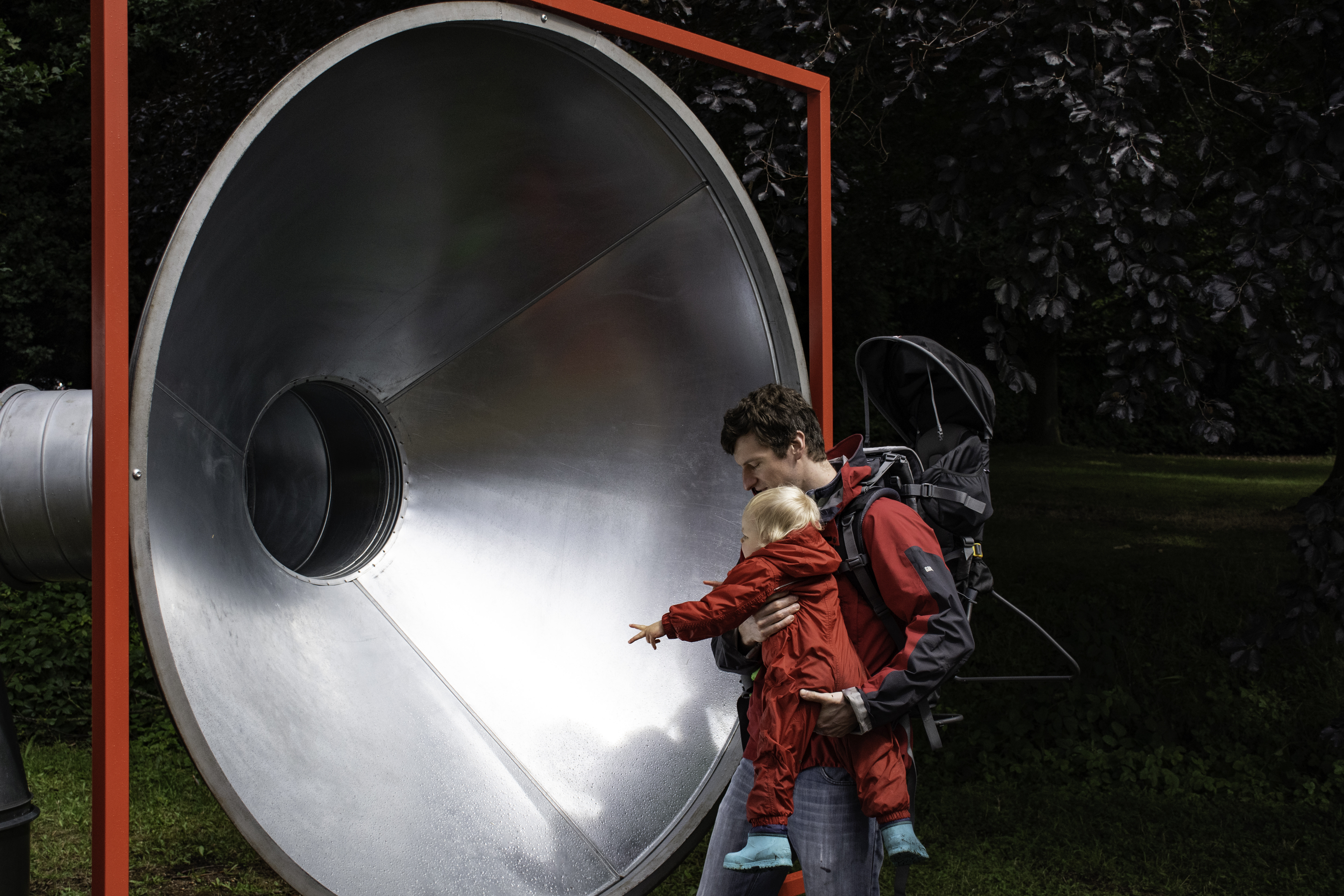 A man and a baby looking into a giant silver tube with a red frame