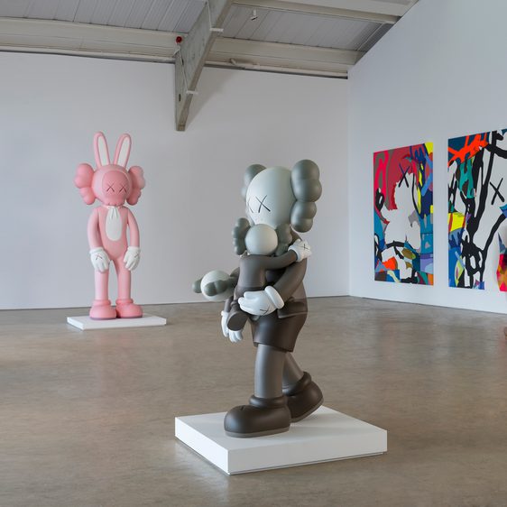 Two large colourful cartoon-style sculptures and wall based artworks in a gallery space. A person can be seen moving between the artworks.