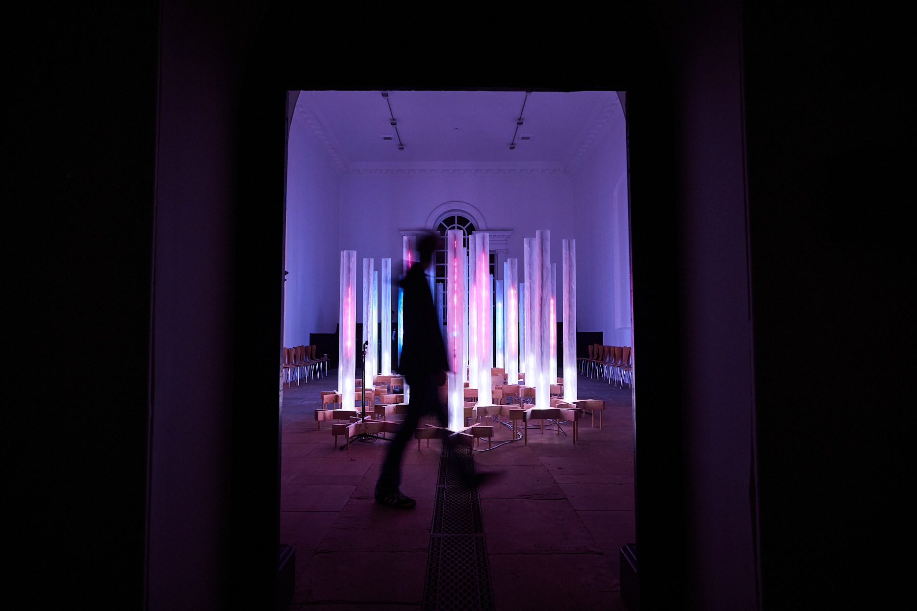 A silhouetted person walking in front of an installation of pink and blue glowing columns.
