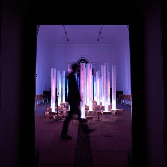 A silhouetted person walking in front of an installation of pink and blue glowing columns.