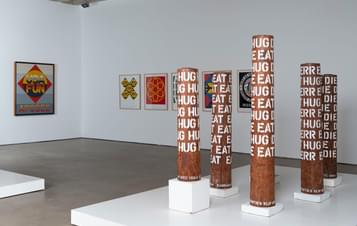 Five pillars painted in text and six paintings hung on gallery wall.