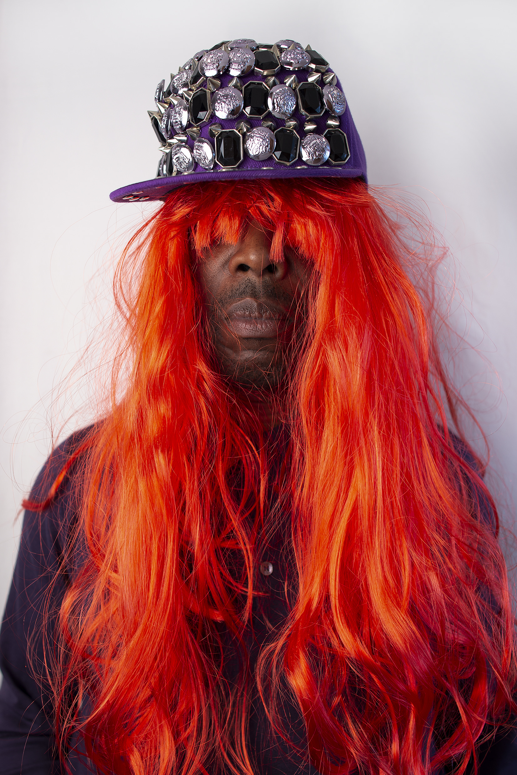 A black man wearing a long red wig and jewel covered baseball cap