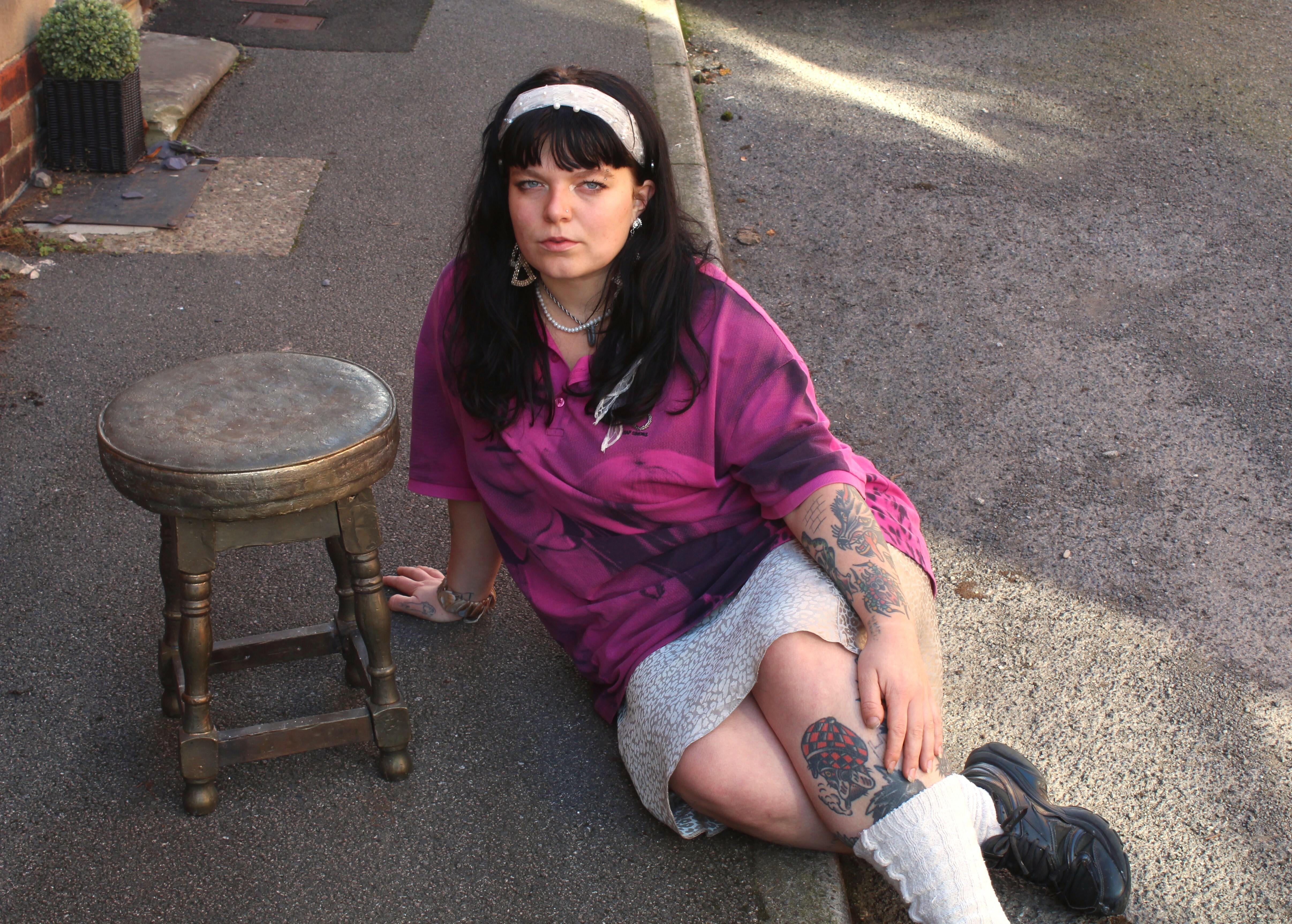 A woman wearing a pink shirt and white skirt, sitting on the ground next to a wooden stool.