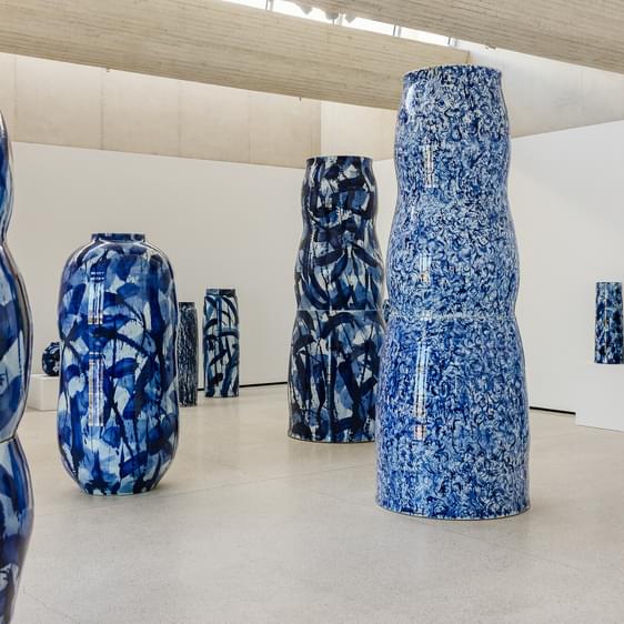 A contemporary art gallery featuring large, abstract blue and white ceramic vases of various shapes and sizes. The vases are arranged throughout the space, with a minimalist white wall and concrete flooring enhancing the display.