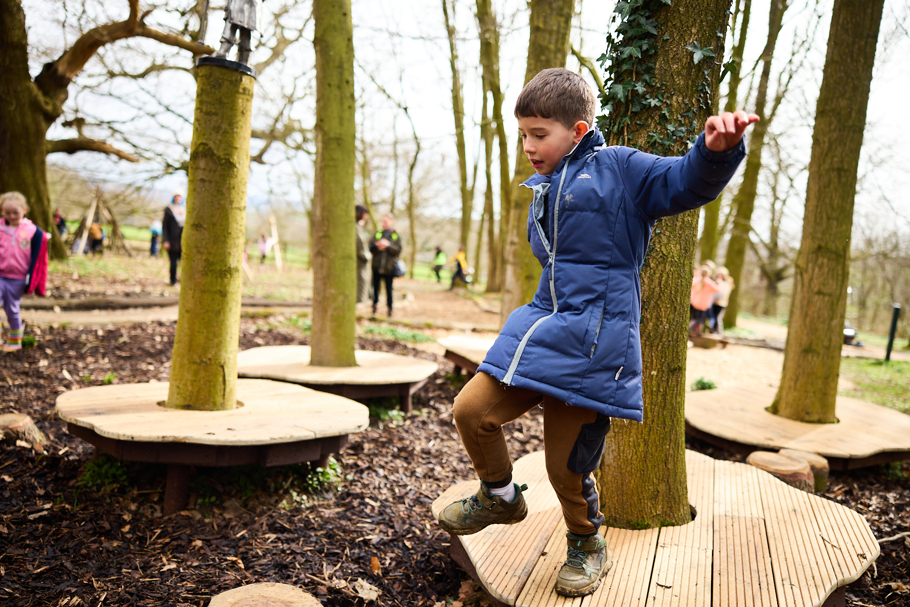 A child jumping from a wooden platform in a woodland play area.