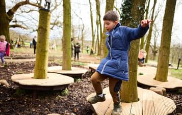 A child jumping from a wooden platform in a woodland play area.