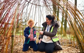 An adult and a child sitting inside a willow den outdoors.