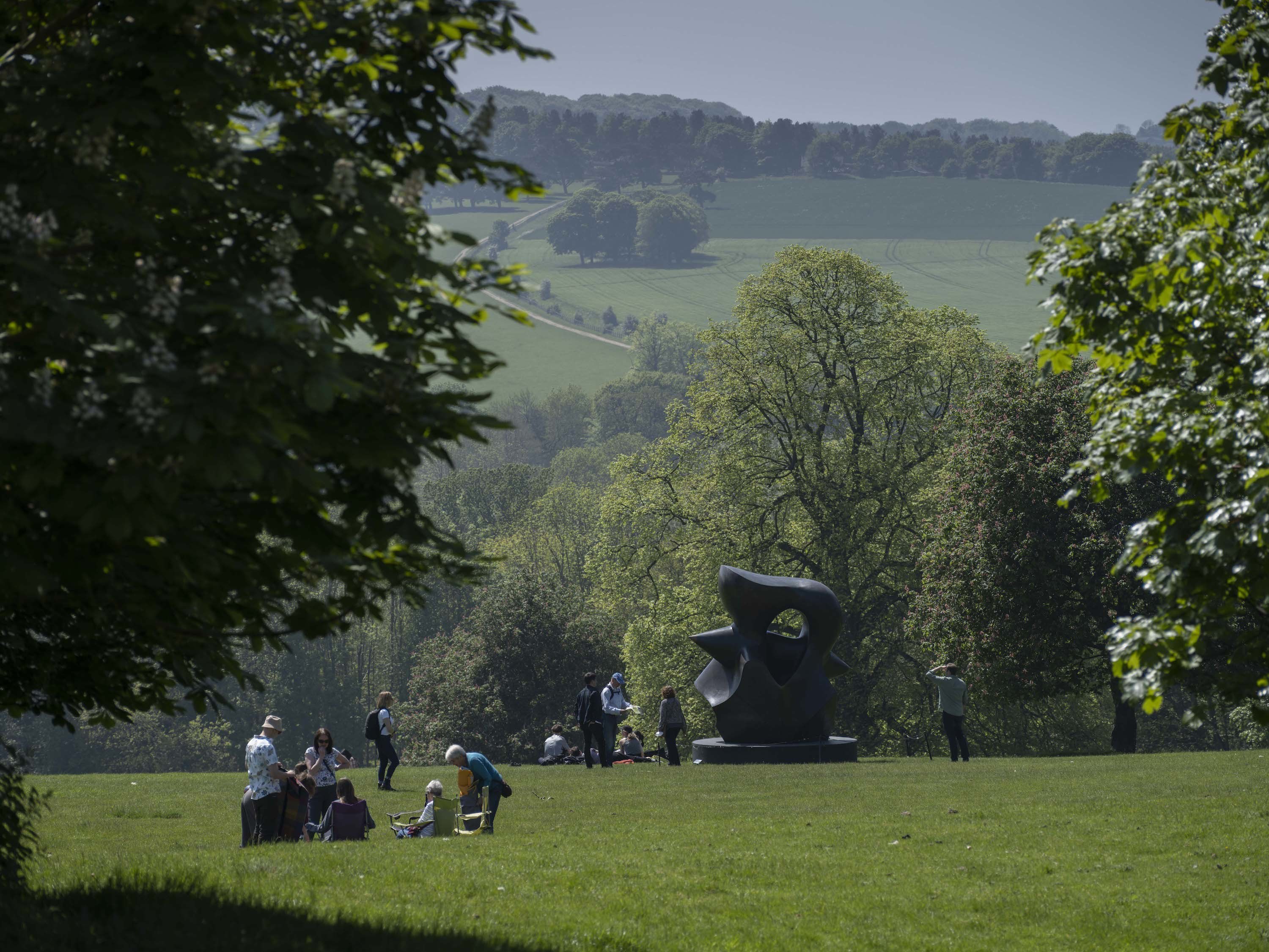 Visitors picnic in chairs whilst other visitors look at a sculpture in the parkland.