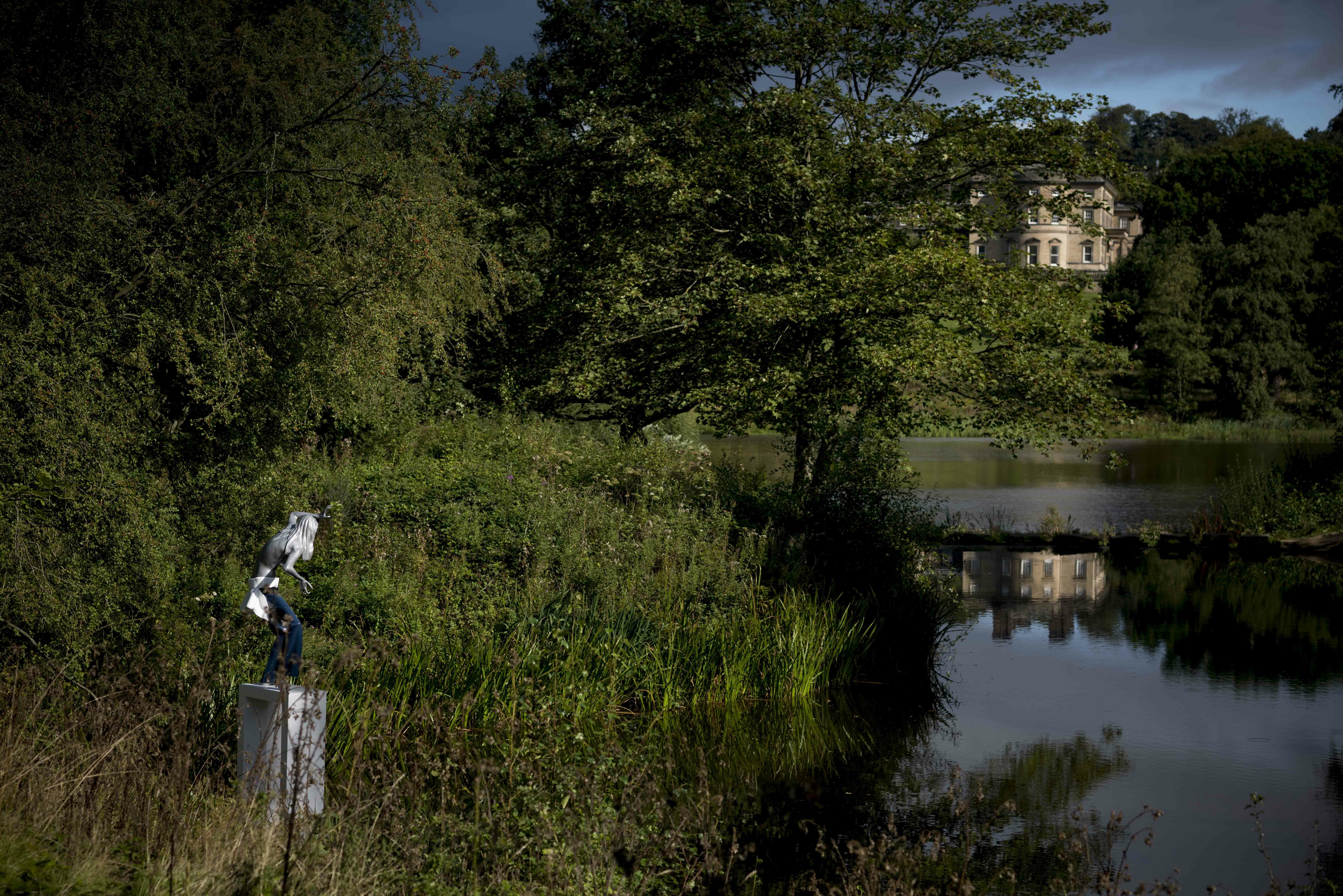 A female figure wearing jeans standing on a white plinth at the edge of a lake, surrounded by foliage. Bretton Hall is in the distance