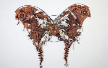 A large, intricate butterfly sculpture made from various materials, including metal and wood. The wings are adorned with gears, feathers, and organic shapes, showcasing a blend of natural and industrial elements against a plain white background.
