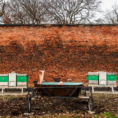 A leg sticking out of a bath tub on wheels in front of a brick wall outdoors between two bee hives.