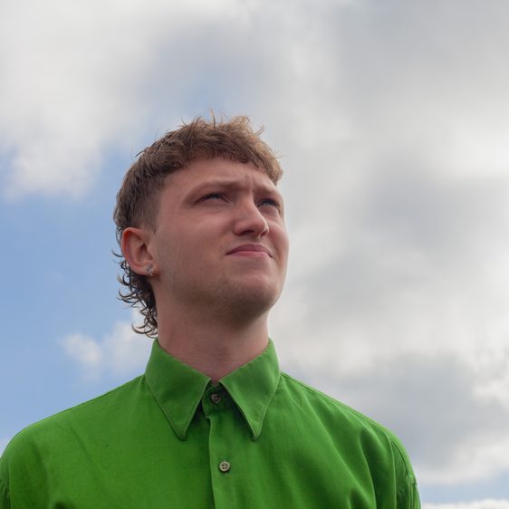 A person with short blonde hair wearing a green shirt in front of a cloudy sky.