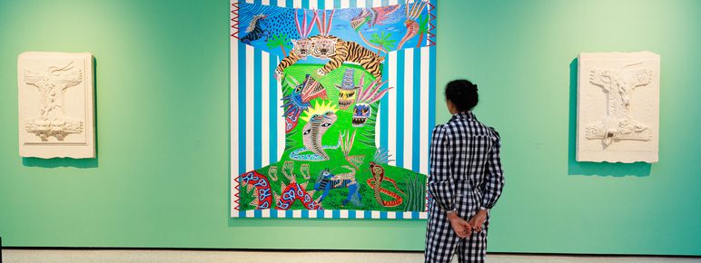 A person in a black and white checked outfit stands in front of a vibrant artwork featuring bold colours and patterns, flanked by two white sculptures on a bright green wall in an art gallery.