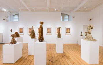 An art gallery featuring several sculptures made from natural materials, displayed on white plinths. The walls are adorned with framed photographs, and the wooden floor adds warmth to the minimalist space, illuminated by natural light.