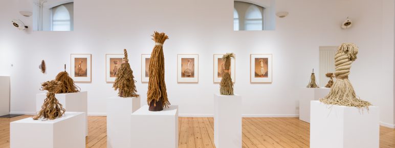 An art gallery featuring several sculptures made from natural materials, displayed on white plinths. The walls are adorned with framed photographs, and the wooden floor adds warmth to the minimalist space, illuminated by natural light.