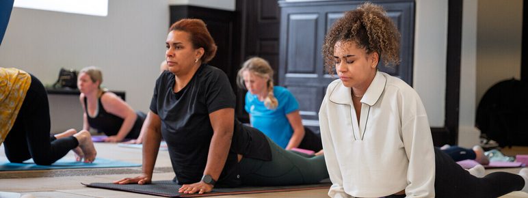A group of people practising yoga in a spacious room. Two women are in the foreground, one in a black top and the other in a white sweatshirt, both in a cobra pose. Others are visible in the background, engaged in various yoga positions.